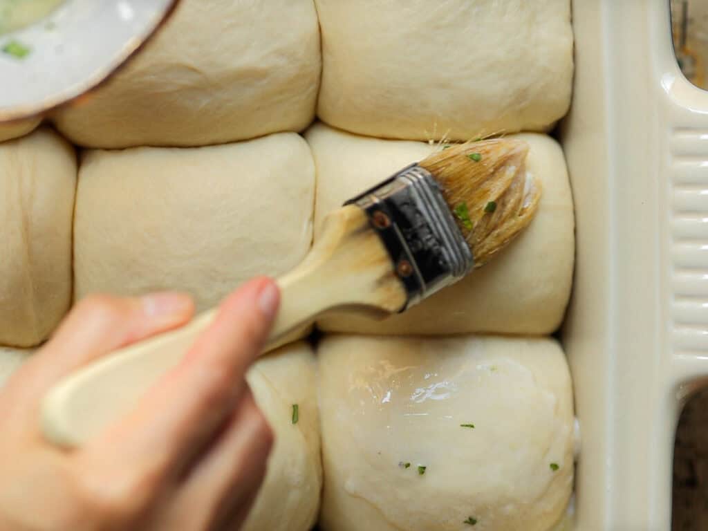 A hand brushes melted butter with herbs onto unbaked dough rolls in a baking dish, preparing them for baking. The rolls are neatly arranged in rows.